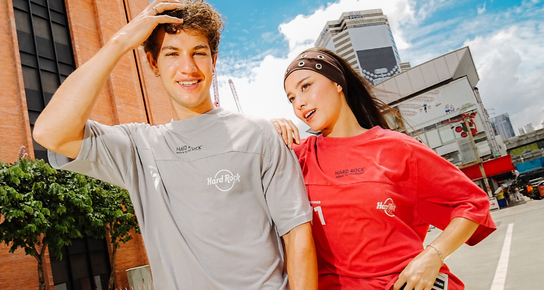 A couple wearing the Hard Rock Move to the Music comfy tees, a man to the left is wearing the said tee in gray, a woman next to him is wearing the said tee in red, both are standing off in the middle of an empty street in a town during the day time. 